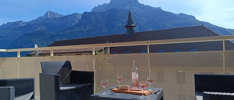 An outdoor seating area with wicker furniture, a table with a bottle and glasses, and a mountain view in the background.