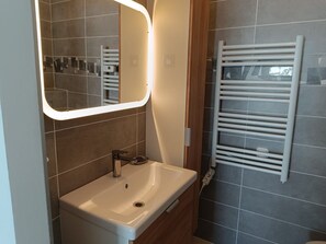 A bathroom with a white sink, a mirror with a light, and a towel rack. - Les Hauts de la Collégiale (4 people) Terrace with a view of Mont Blanc & free parking. (Sallanches)
