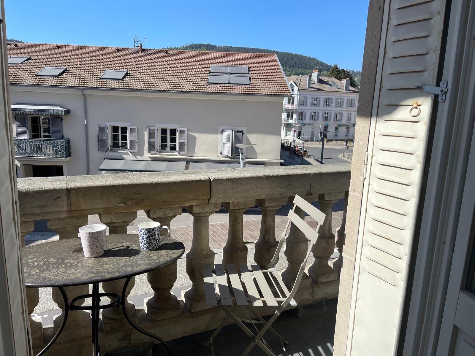 Un balcon avec une table et des chaises, donnant sur une rue bordée d’immeubles et sur une colline.