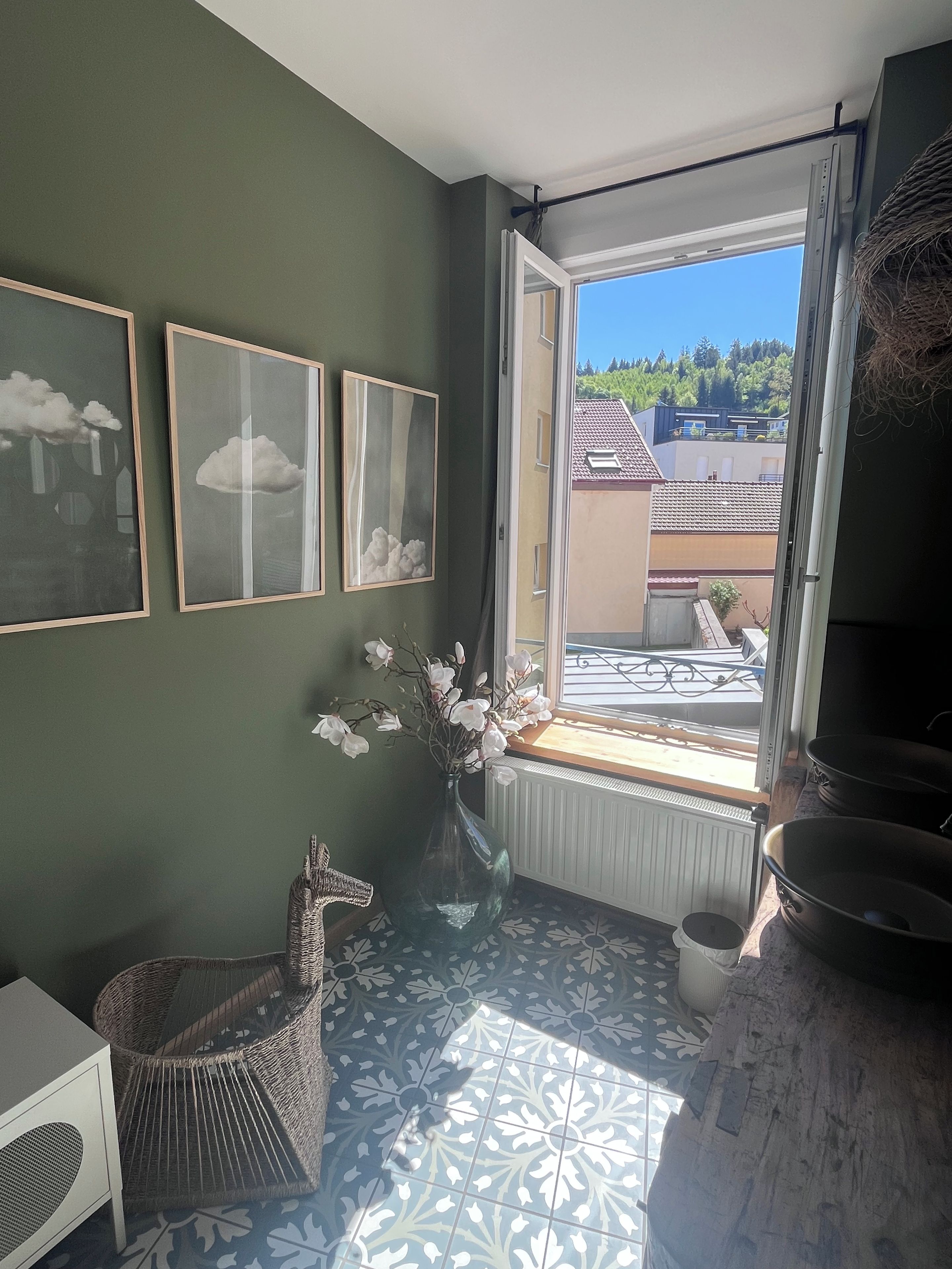 Een kamer met uitzicht op een woonwijk, een rieten stoel, een vaas met bloemen en ingelijste foto's aan de muur.