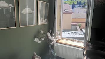 Een kamer met uitzicht op een woonwijk, een rieten stoel, een vaas met bloemen en ingelijste foto's aan de muur.