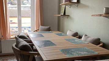 A dining area with a wooden table and chairs, a view through a window, and a wall with shelves.
