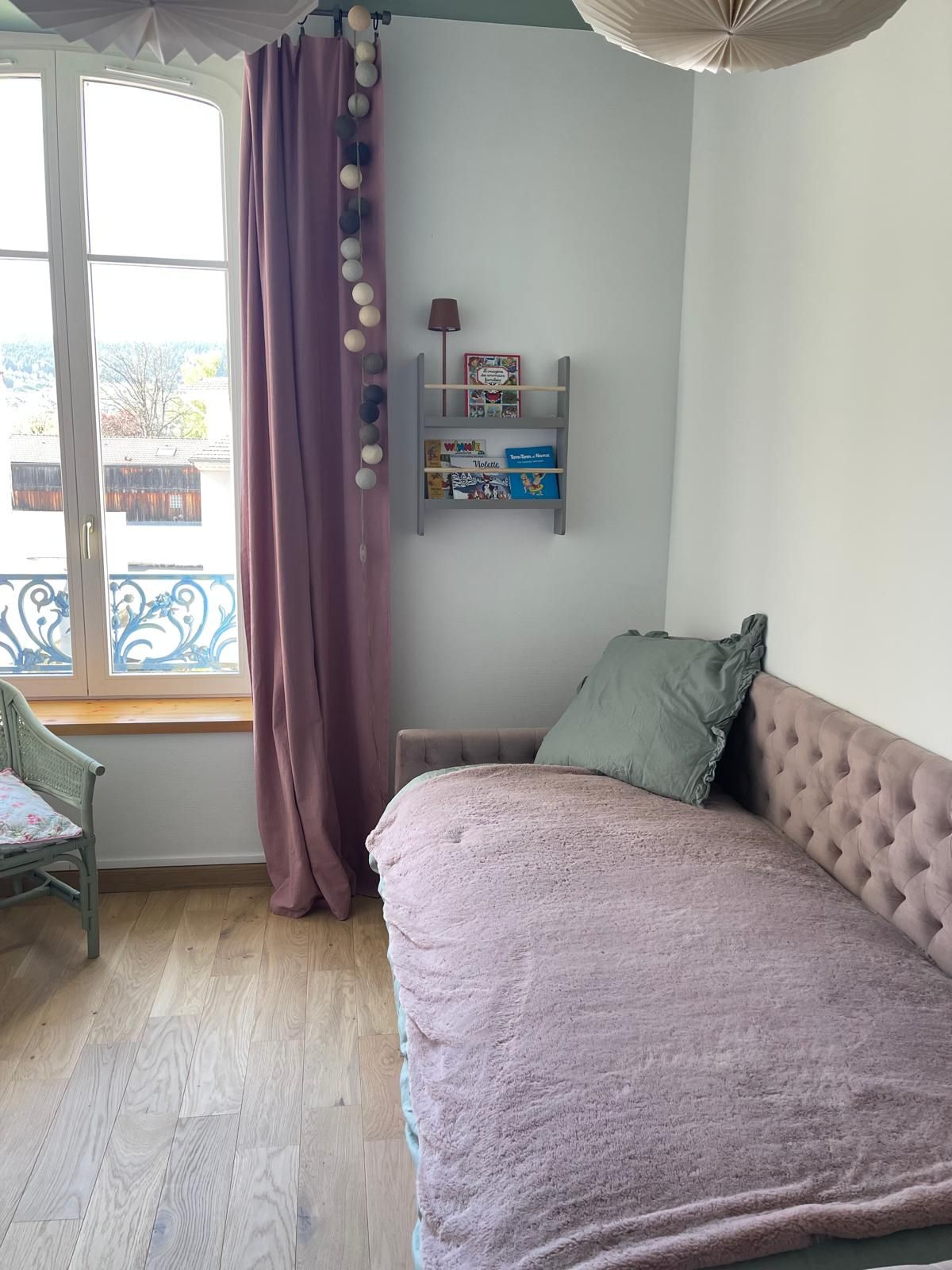 A bedroom with a bed, a window with curtains, a chair, and a shelf with books.