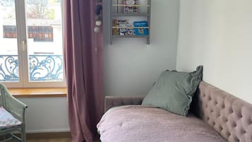 A bedroom with a bed, a window with curtains, a chair, and a shelf with books.