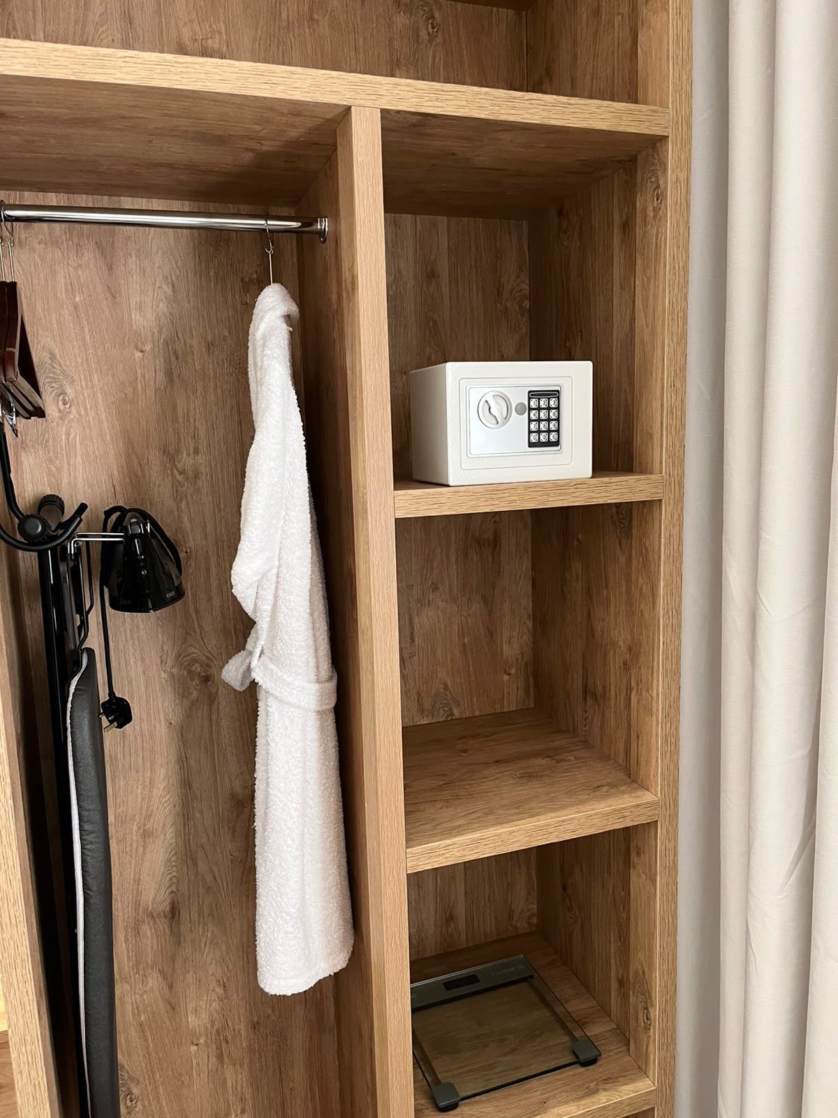A wooden shelf unit with a white robe hanging on a hook, a white electronic device, and a black object on the lower shelf.