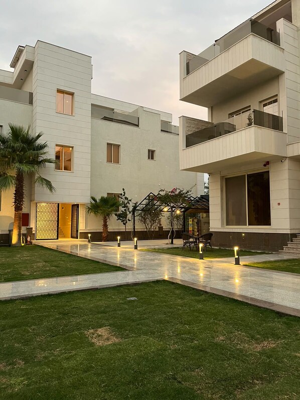 A modern building with a flat roof, multiple balconies, and a well-maintained courtyard with a palm tree and pathway lighting.