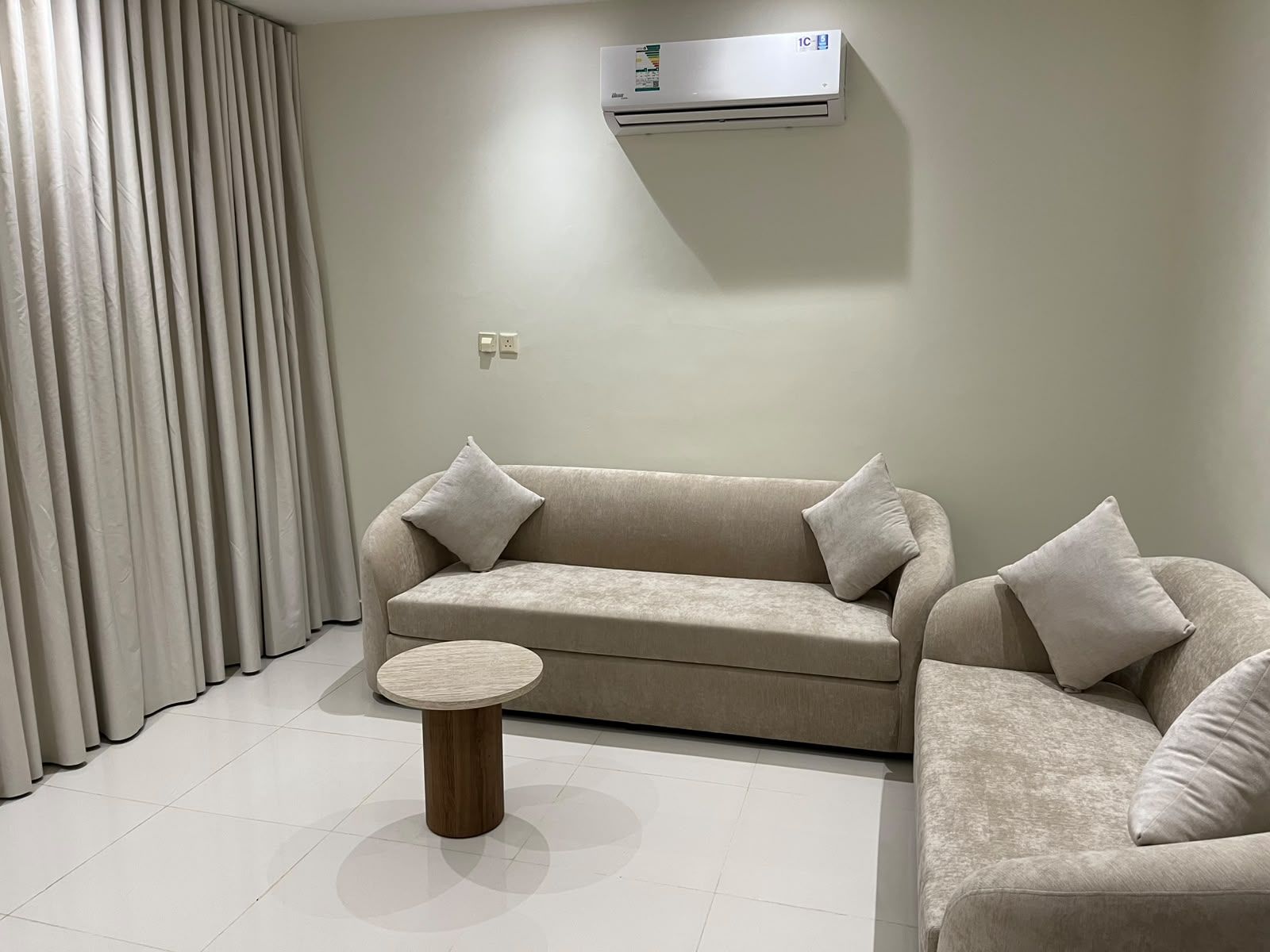 A living room with a beige sofa, two pillows, a round wooden coffee table, and an air conditioning unit mounted on the wall.
