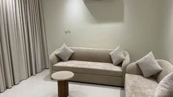 Ein Wohnzimmer mit einem beigefarbenen Sofa, zwei Kissen, einem runden Holztisch und einer an der Wand montierten Klimaanlage.