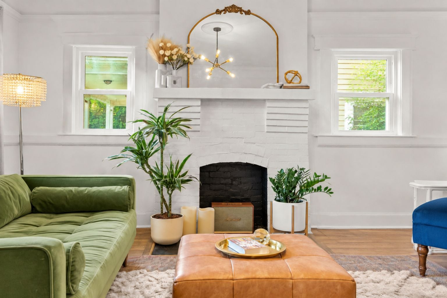 A living room with a green sofa, a brown ottoman, a fireplace, and two windows with natural light.
