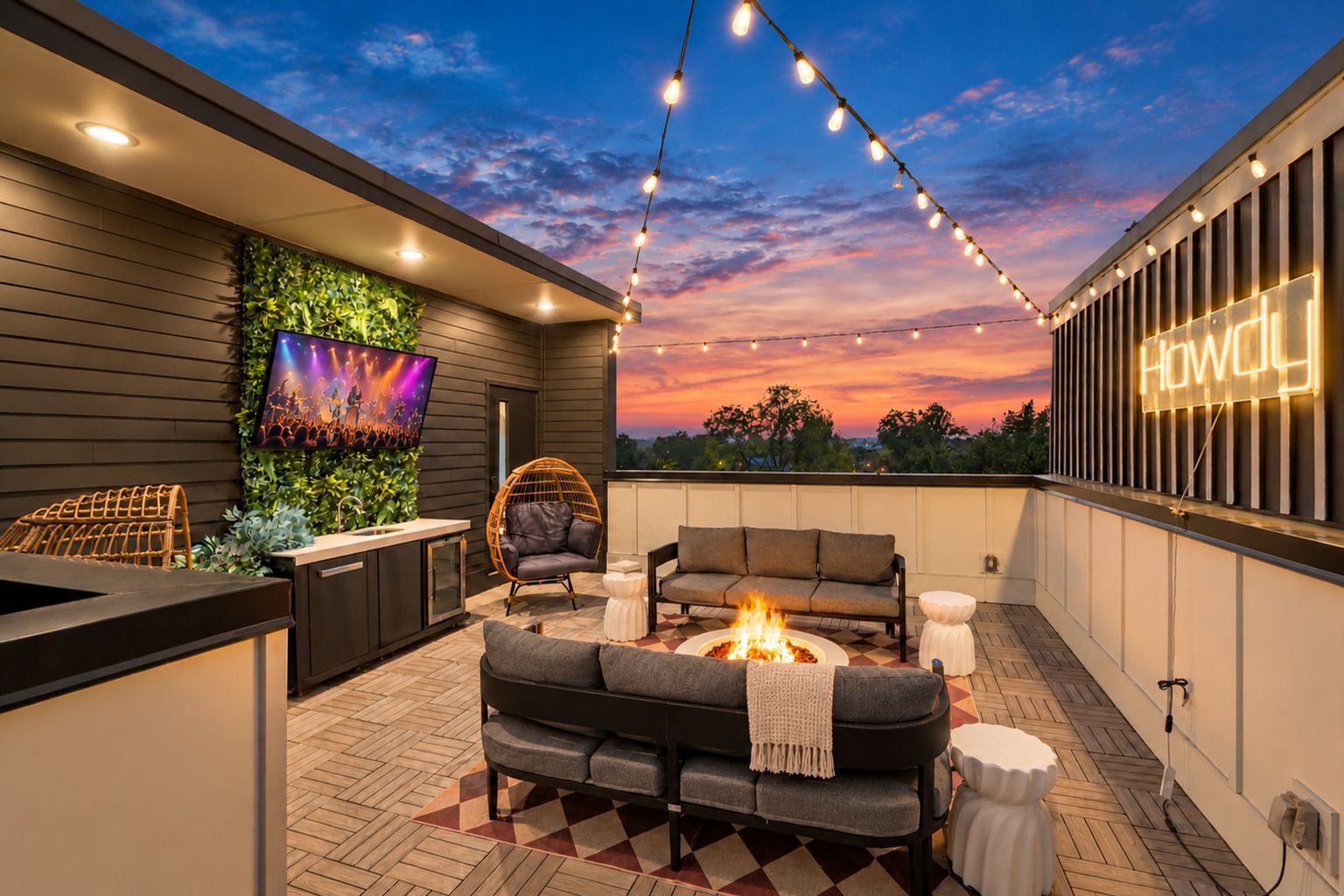 An outdoor patio with a fire pit, seating, a TV, and string lights against a sunset sky.