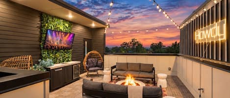 Un patio al aire libre con un brasero, asientos, un televisor y luces colgantes bajo un cielo al atardecer.