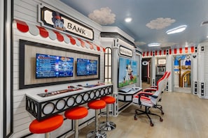 A themed bar area with a 'Bank' sign, multiple screens displaying games, red bar stools, and a Mickey Mouse mural. - Brand NEW 5BR w Pool Themed Rooms Lazy River 16220 (Clermont)