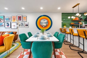 A dining area with a white table set for four, surrounded by teal chairs. There is a colorful wall art piece above the table and a kitchen area with a green backsplash and yellow chairs. - Walkable Sobro House - Gated Parking - Sleeps 8 (Nashville)