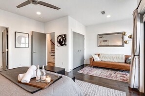 A modern hotel room with a bed, a wooden side table with bottles and a candle, a brown leather sofa, a patterned rug, and a large mirror on the wall. - Gulch Rooftop Fire pit Putting Green - Sleeps 12 (Nashville)