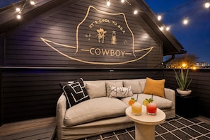 A dark wooden wall with a neon sign that reads 'IT'S COOL TO BE A COWBOY' and a sofa with cushions and a small table with drinks. - Rooftop Patio Home Near Broadway Vandy - Sleeps 12 (Nashville)