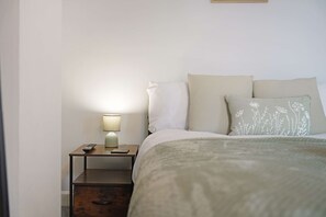 Een opgemaakt bed met een deken met wit en groene patronen, een bedlampje en een afstandsbediening op een houten nachtkastje.