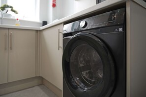 Vooraanzicht van een moderne wasmachine in een keuken met een wit aanrechtblad en beige kastjes.