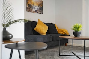 Een moderne woonkamer met een donkere bank, een ronde houten tafel en een potplant.
