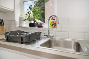 A kitchen with a stainless steel sink, a dish rack, a potted plant, and a window with a view of greenery. - Cozy 2BR Home Sleeps 5 With Wifi by Stay With Us (Wigan)