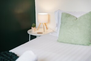 Ein ordentlich bezogenes Bett mit einem grünen Kopfkissen, ein Nachttisch mit einer Lampe und einer kleinen Pflanze.