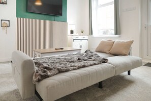 Ein modernes Wohnzimmer mit einer Couch, einem an der Wand montierten Fernseher, einer Küchenzeile und einem Fenster mit Vorhängen.