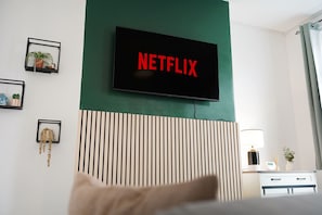 A hotel room with a television mounted on a green wall displaying the Netflix logo, a bed with a pillow, a bedside table with a lamp, and a small plant on the nightstand. - Cosy Blackhall 2BR Sleeps 5 By Stay With Us (Hartlepool)