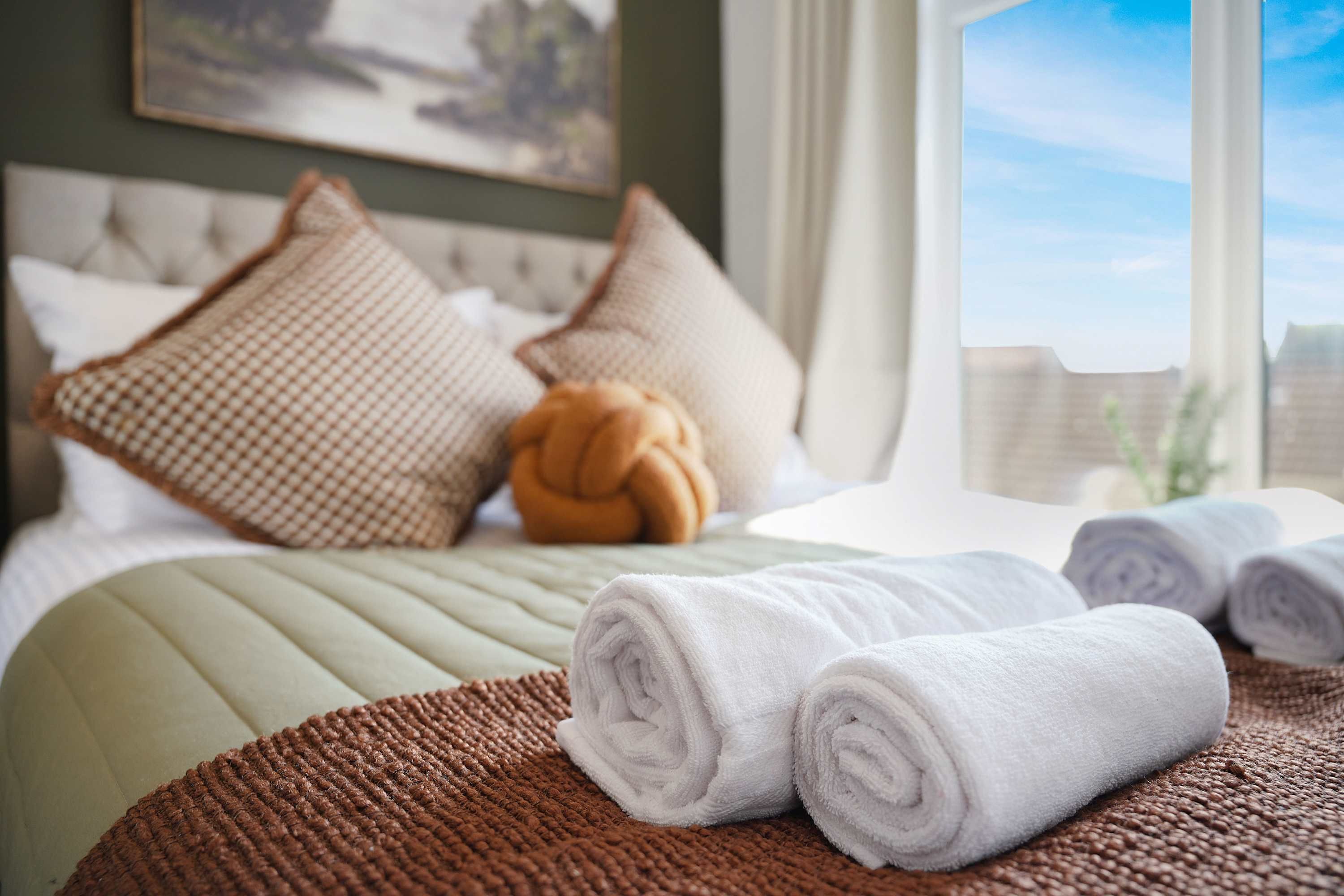 Una cama bien hecha con toallas blancas enrolladas, una manta marrón texturizada y almohadas decorativas.