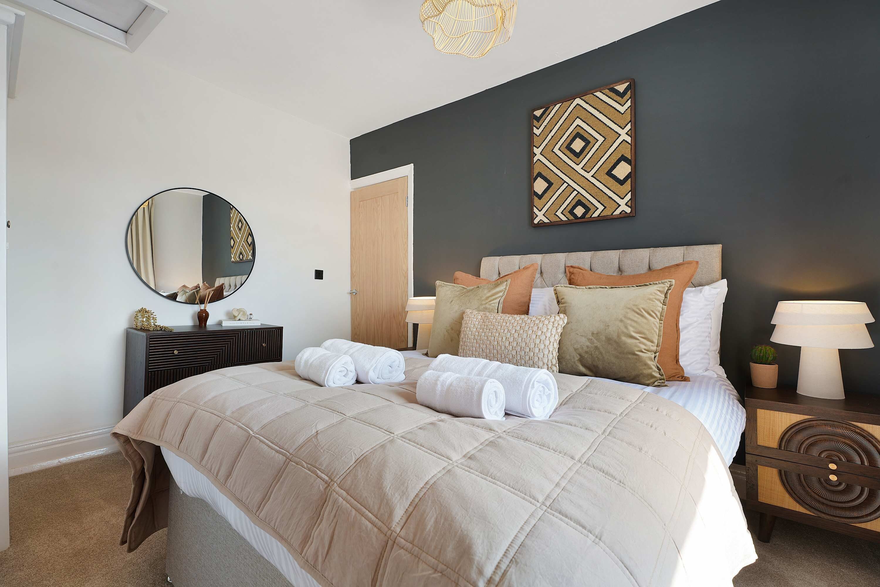 Una cama bien hecha con colcha color beige, una mesita de noche con lámpara y un espejo montado en la pared.