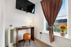Ein Zimmer mit einem an der Wand befestigten Flachbildfernseher, einem Schreibtisch mit Lampe, einem braunen Lederhocker, einem Heizkörper und einem Fenster mit Vorhängen.