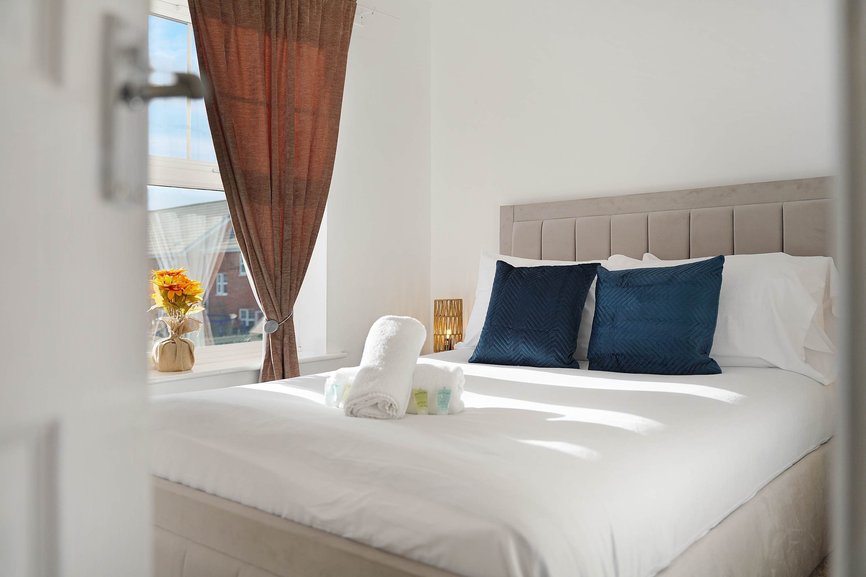 Una cama bien hecha con almohadas y una toalla enrollada, una ventana con cortinas y un jarrón con flores en el alféizar.