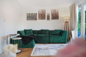 Ein Wohnzimmer mit einem grünen Sofa, einem kleinen Beistelltisch, einer Stehlampe und drei an der Wand hängenden Bildern.