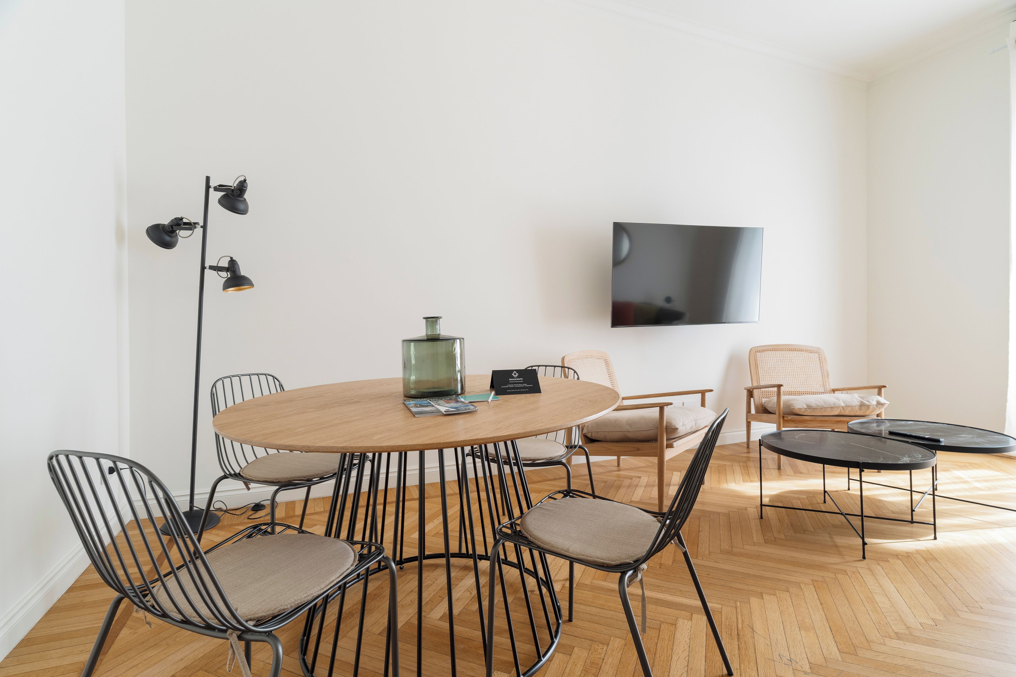 Een moderne woonkamer met een ronde houten tafel, zwarte metalen stoelen, een flatscreen televisie en een vloerlamp.