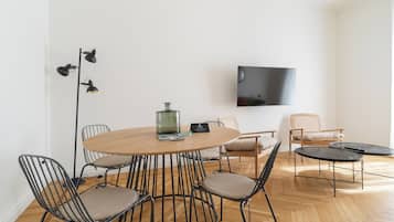 Een moderne woonkamer met een ronde houten tafel, zwarte metalen stoelen, een flatscreen televisie en een vloerlamp.