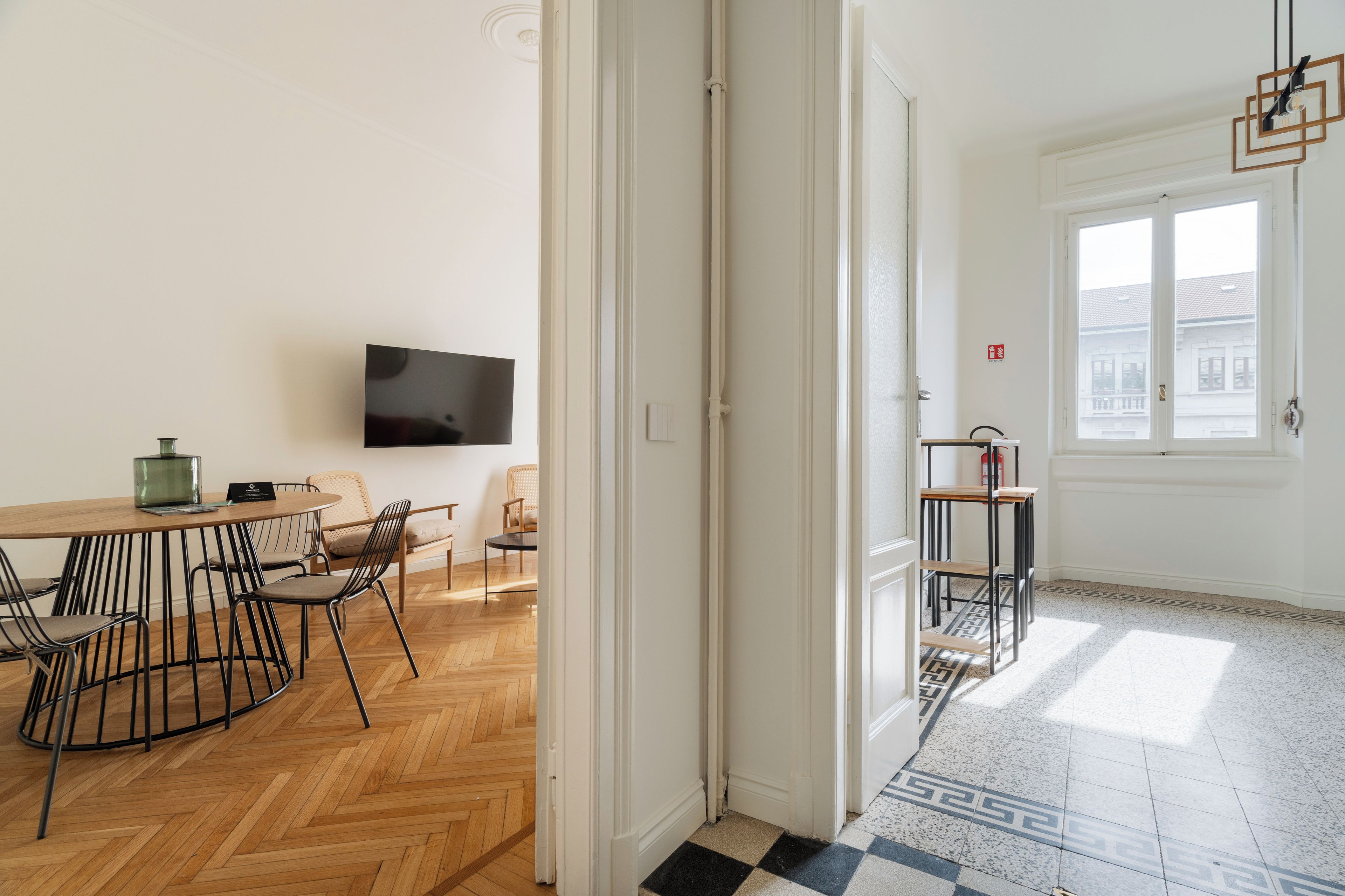 Een kamer met een houten visgraatvloer, een eettafel met stoelen, een flatscreen televisie aan de muur en een raam met uitzicht op gebouwen.