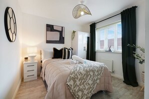 A bedroom with a bed, bedside table, lamp, abstract art, window with curtains, and a radiator. - Modern 4BR Stay Driveway Parking By Stay With Us (Middlesbrough)