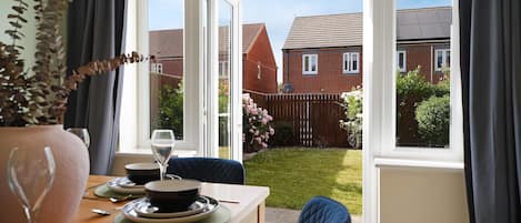 A dining area with a table set for two, a vase with dried flowers, and a view of a residential neighborhood through large windows.