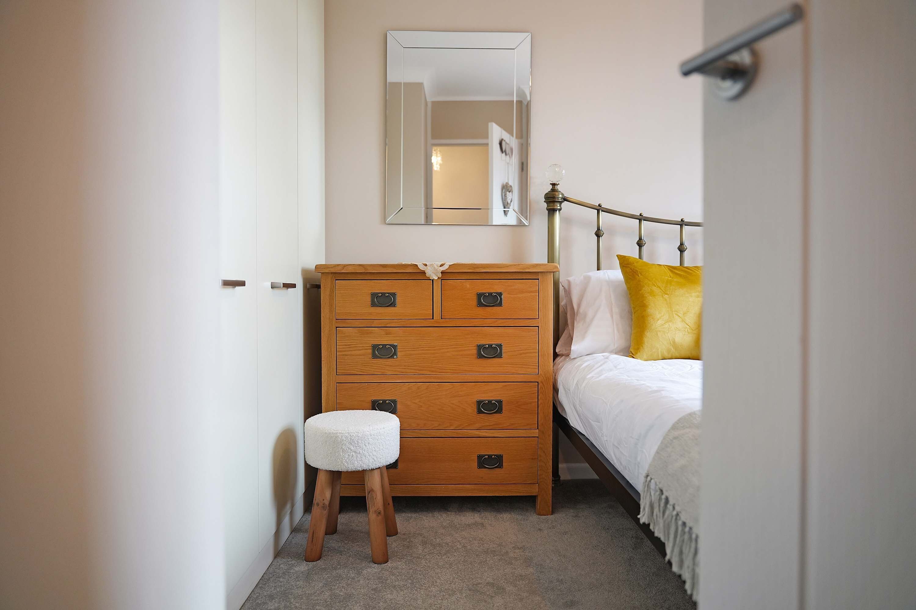 Ein Schlafzimmer mit einem hölzernen Schminktisch, einem Einzelbett mit Metallkopfteil, einem kleinen Hocker und einem Spiegel an der Wand.
