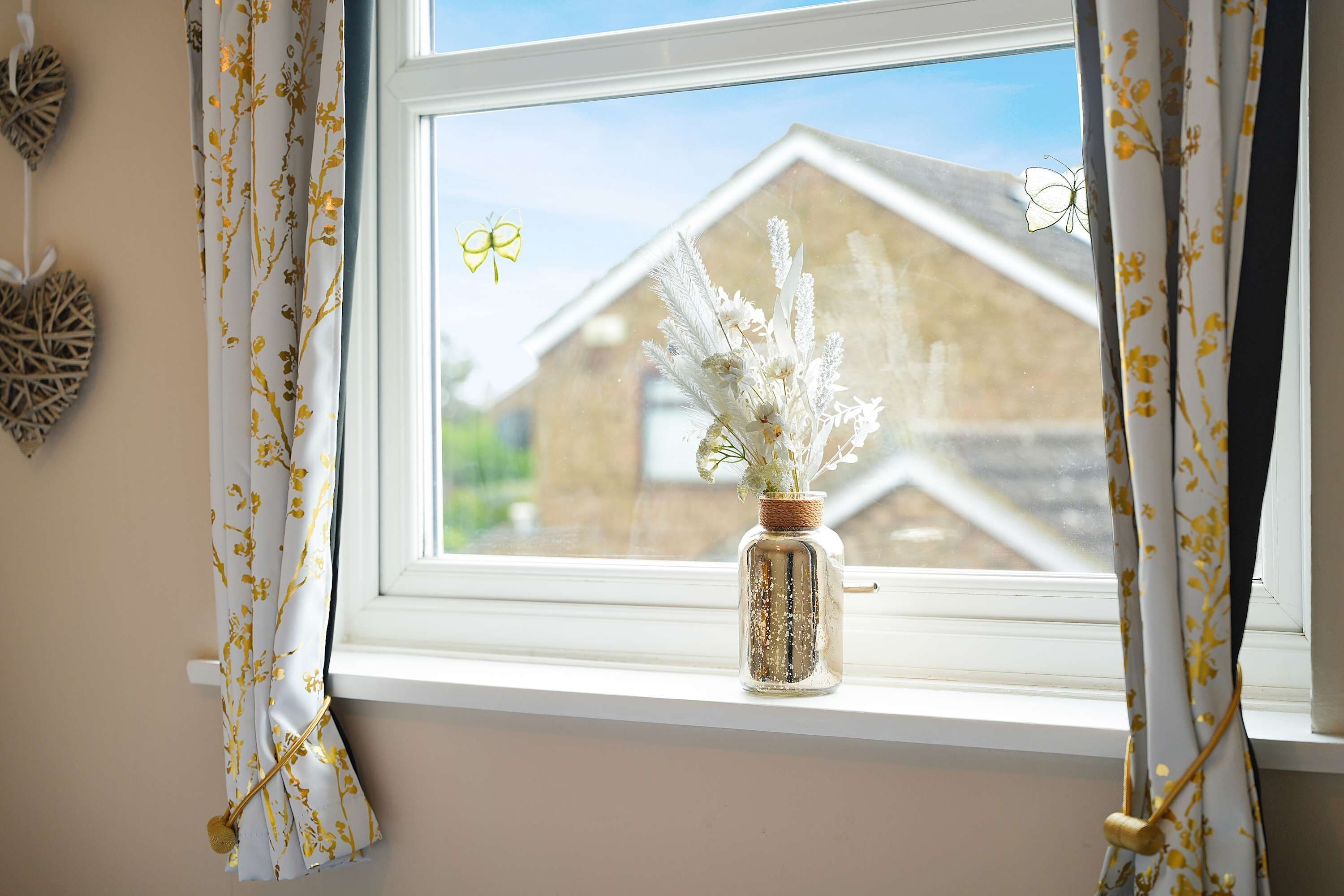 A window with a view of a house, curtains with a floral pattern, and a vase with white flowers on the windowsill.