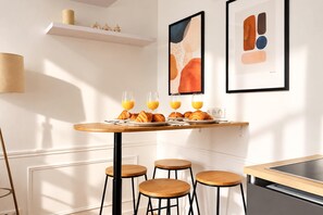 Desayuno con cruasanes y jugo de naranja sobre una mesa de madera, con taburetes y una pared blanca decorada con arte.