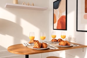 Ein Frühstückstisch mit Croissants, Orangensaft und einer Tischplatte in einem hellen Zimmer mit weißer Wand und einem Regal mit Dekoration.