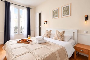 Quarto de hotel com cama, mesa de cabeceira, bandeja com croissants e suco de laranja, janela com cortinas e duas luminárias fixadas na parede.