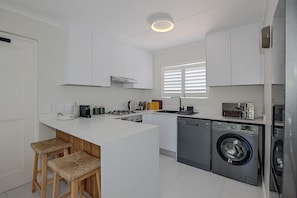 A modern kitchen with white cabinetry, a central island with stools, a built-in washing machine, and a window with blinds. - Stylish 2-bedroom apartment in enchanting Big Bay neighborhood (Cape Town)
