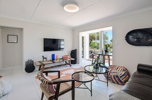 A modern living room with a flat-screen TV, a dining area with a wooden table and chairs, a sofa, and a large window offering a view of the outdoors. - Stylish 2-bedroom apartment in enchanting Big Bay neighborhood (Cape Town)