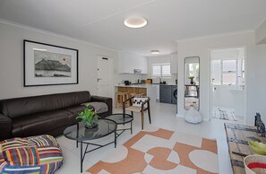Uma sala de estar moderna com um sofá de couro marrom, uma mesa de centro de vidro e uma almofada colorida. Uma cozinha com armários brancos e uma área de jantar com mesa e cadeiras de madeira são visíveis ao fundo.