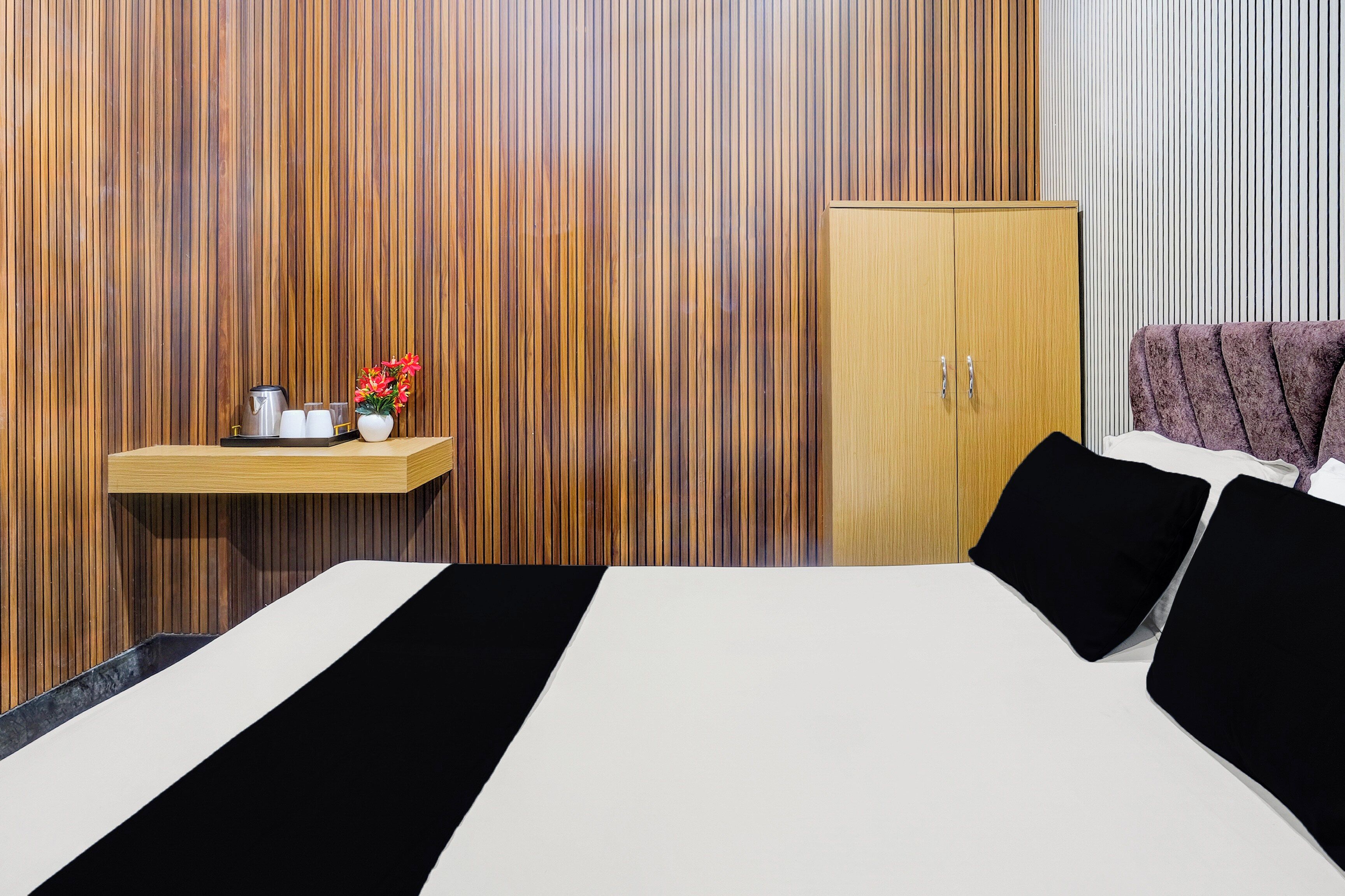 Une chambre d’hôtel moderne avec un mur lambrissé en bois, un lit avec une literie noire et blanche, une petite étagère en bois contenant des articles de toilette et une tête de lit violette.
