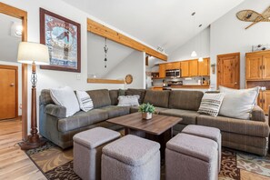 Un salon moderne avec un canapé d’angle, des poufs, une table basse et une cuisine en arrière-plan.