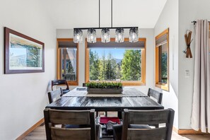 Une salle à manger avec une grande table, des chaises, un lustre et une vue sur les arbres à travers de grandes fenêtres.