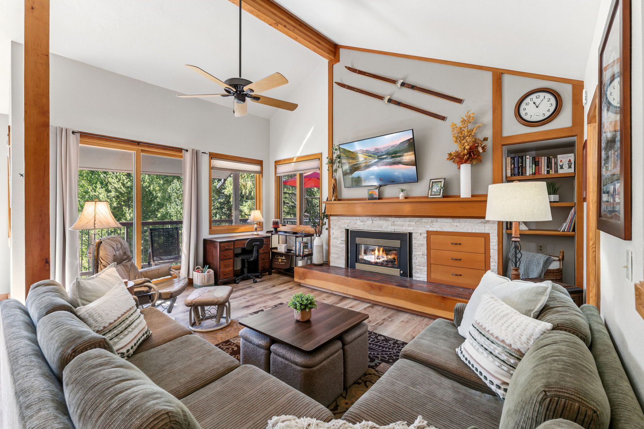 A spacious living room with a fireplace, a television mounted above it, and a large window providing a view of the outdoors.