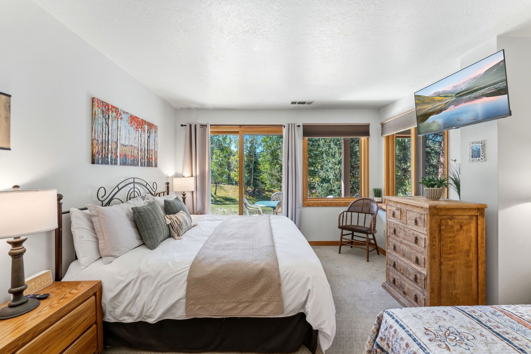 Quarto com cama, mesa de cabeceira, abajur, cômoda, cadeira e uma janela grande com vista para árvores.