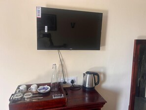 A flat-screen TV mounted on a wall above a wooden table with a coffee maker, cups, and a glass pitcher. - Marwar leopard Camp jawai (Bali)